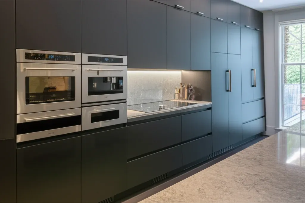 Long handleless kitchen run in dark teal-grey with two stainless wall-oven stacks and full-height panelled cabinetry beside them